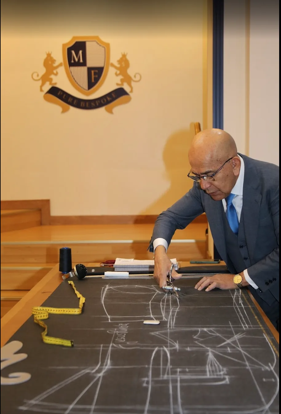 Manuel Fernández trazando a tiza sobre el tejido en la mesa de corte, bajo el escudo de la casa